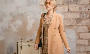 Elegancka kobieta w płaszczu w kolorze chamois, z kwiecistą sukienką i okularami przeciwsłonecznymi, pozuje przy starej walizce i ceglanej ścianie.