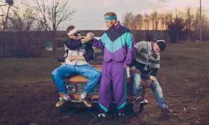 Trzech mężczyzn w strojach z lat 90. moda, siedzących na starym samochodzie. Jeden w dresie, drugi w bluzie i jeansach.
