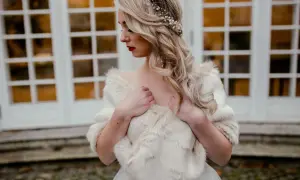 Elegancka panna młoda w białej etoli z futra, ozdobiona perłami i złotymi elementami we włosach, pozuje przed drzwiami.