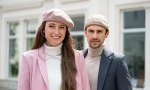 Uśmiechnięta kobieta w różowym żakiecie i beretce, obok mężczyzna w beżowym golfie i marynarce. Stylowa dokerka na głowie kobiety.