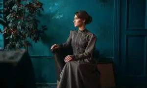 Elegancka kobieta w sukni w stylu dark academia siedzi w fotelu obok rośliny.
