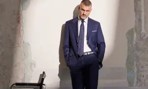 Elegancki mężczyzna w granatowym garniturze, z dłońmi w kieszeniach, stoi przy zniszczonej ścianie.
