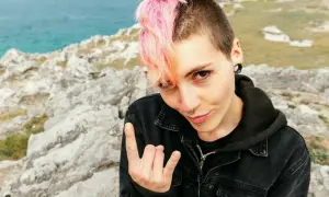 Dziewczyna z różowymi włosami i kolczykami, w kurtce dżinsowej, pozuje na tle skał i morza, pokazując znak rocka. Jej styl to czysty **punk**.