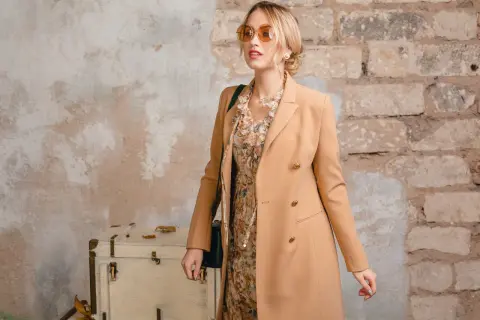 Elegancka kobieta w płaszczu w kolorze chamois, z kwiecistą sukienką i okularami przeciwsłonecznymi, pozuje przy starej walizce i ceglanej ścianie.