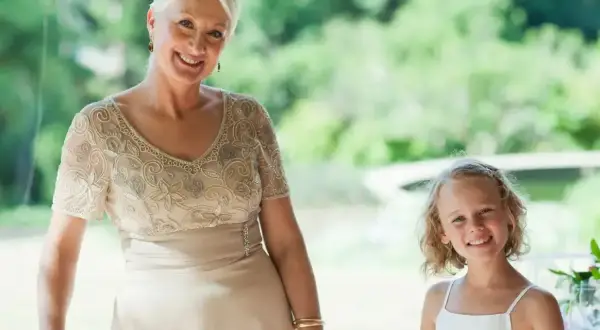 Elegancka stylizacja na komunię dla babci. Uśmiechnięta kobieta w beżowej sukni z haftem i dziewczynka w bieli.