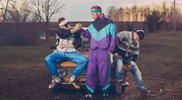 Trzech mężczyzn w strojach z lat 90. moda, siedzących na starym samochodzie. Jeden w dresie, drugi w bluzie i jeansach.