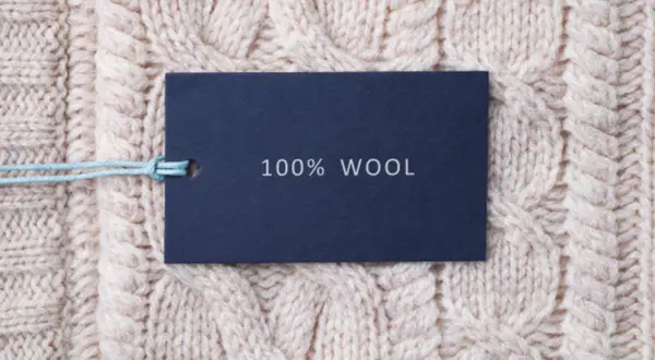 Metka z napisem "100% WOOL" na tle grubego, beżowego swetra w warkocze. To właśnie jest lambswool – czysta wełna.
