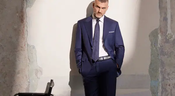 Elegancki mężczyzna w granatowym garniturze, z dłońmi w kieszeniach, stoi przy zniszczonej ścianie.
