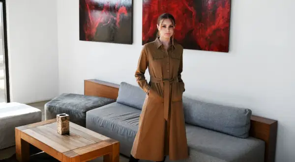 Kobieta w brązowej, casualowej sukience z paskiem stoi w nowoczesnym salonie.