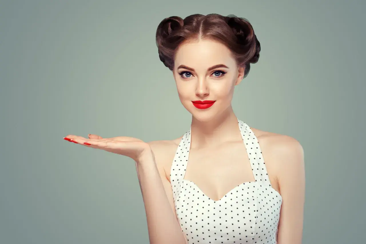 Kobieta w stylu pin-up z burzą loków i czerwoną szminką prezentuje coś na otwartej dłoni.