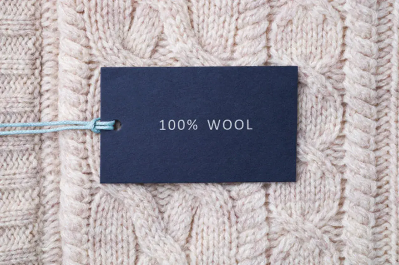 Metka z napisem "100% WOOL" na tle grubego, beżowego swetra w warkocze. To właśnie jest lambswool – czysta wełna.