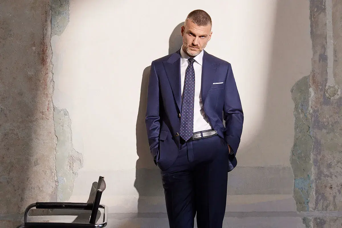 Elegancki mężczyzna w granatowym garniturze, z dłońmi w kieszeniach, stoi przy zniszczonej ścianie.