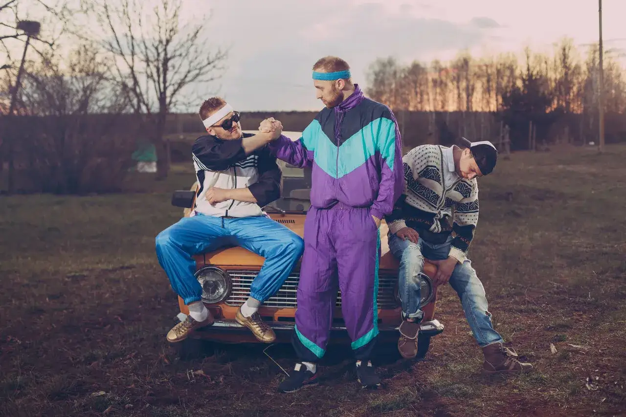 Trzech mężczyzn w strojach z lat 90. moda, siedzących na starym samochodzie. Jeden w dresie, drugi w bluzie i jeansach.