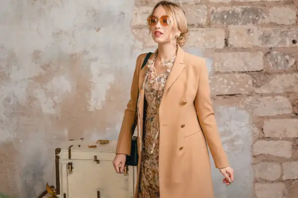 Elegancka kobieta w płaszczu w kolorze chamois, z kwiecistą sukienką i okularami przeciwsłonecznymi, pozuje przy starej walizce i ceglanej ścianie.
