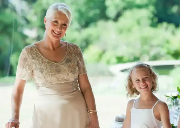 Elegancka stylizacja na komunię dla babci. Uśmiechnięta kobieta w beżowej sukni z haftem i dziewczynka w bieli.