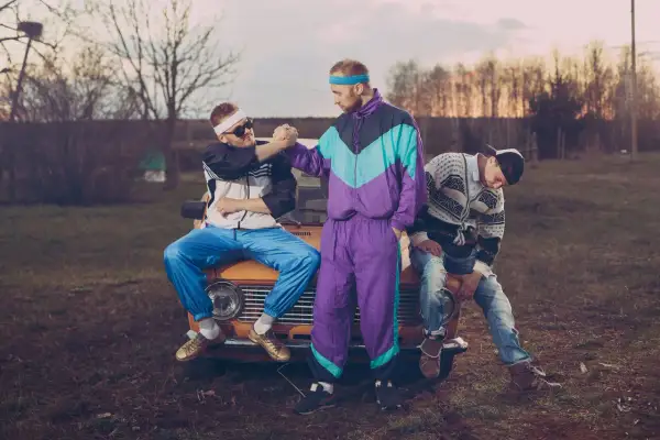 Trzech mężczyzn w strojach z lat 90. moda, siedzących na starym samochodzie. Jeden w dresie, drugi w bluzie i jeansach.