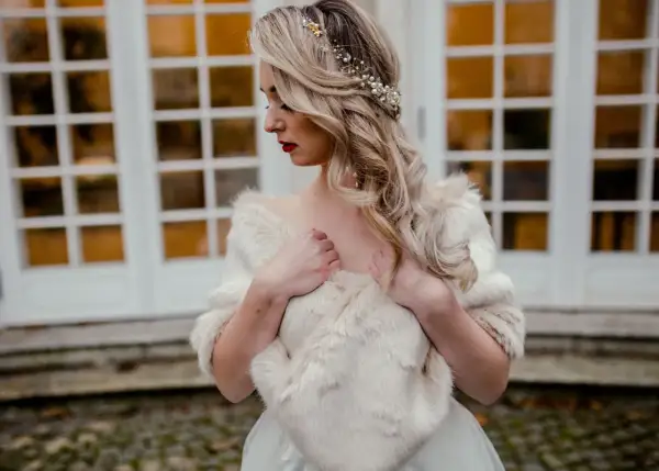 Elegancka panna młoda w białej etoli z futra, ozdobiona perłami i złotymi elementami we włosach, pozuje przed drzwiami.