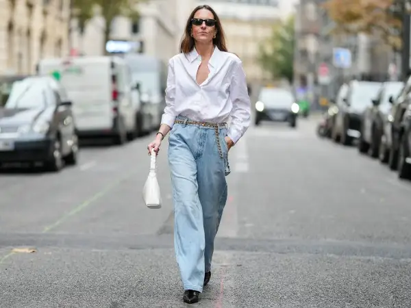 Kobieta w luźnych, jasnych jeansach typu mom fit i białej koszuli, z białą torebką. Idealny look dla każdej kobiety ceniącej wygodę i styl.