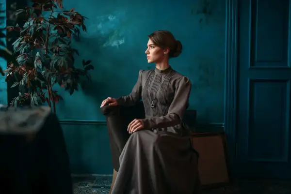 Elegancka kobieta w sukni w stylu dark academia siedzi w fotelu obok rośliny.