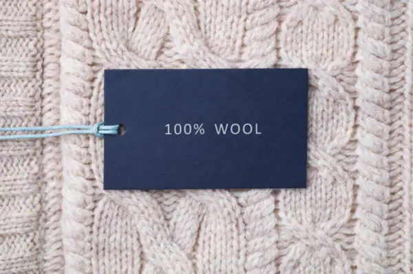 Metka z napisem "100% WOOL" na tle grubego, beżowego swetra w warkocze. To właśnie jest lambswool – czysta wełna.