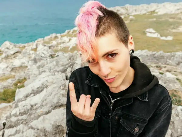 Dziewczyna z różowymi włosami i kolczykami, w kurtce dżinsowej, pozuje na tle skał i morza, pokazując znak rocka. Jej styl to czysty **punk**.